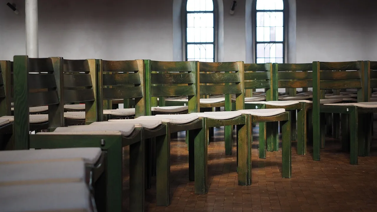 Des rangées de chaises en bois vert dans une salle aux hautes fenêtres